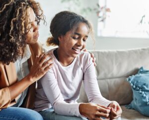 A parent gently places a hand on their child’s shoulder while they talk and smile together on a cozy living room couch, showing support and connection.