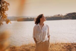 Woman standing by a lake with her eyes closed, enjoying the gentle breeze and warm sunlight in a peaceful outdoor setting.